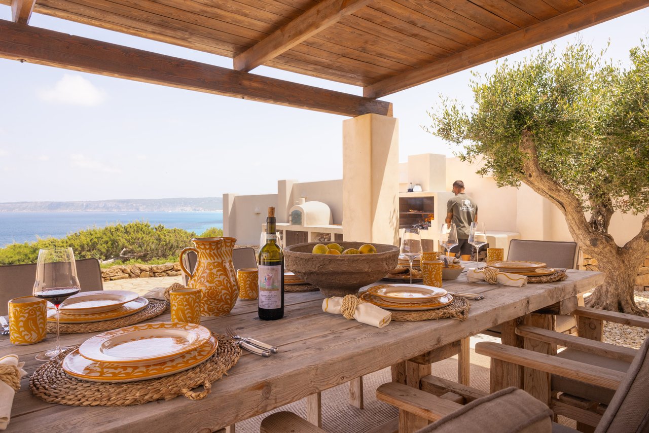 Mesa puesta bajo la pérgola con vajilla amarilla y vino, frente al mar