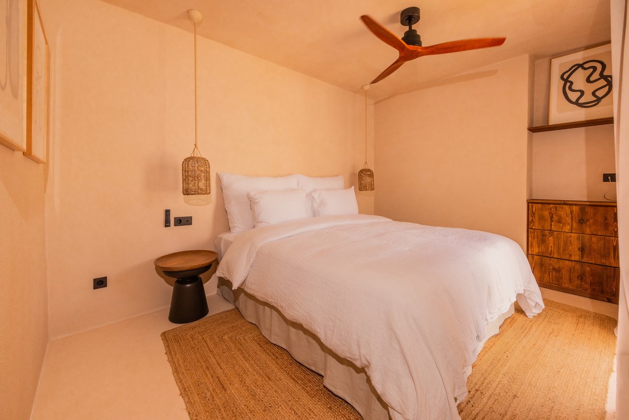 Habitación 04: cama blanca, cómoda de madera y ventilador